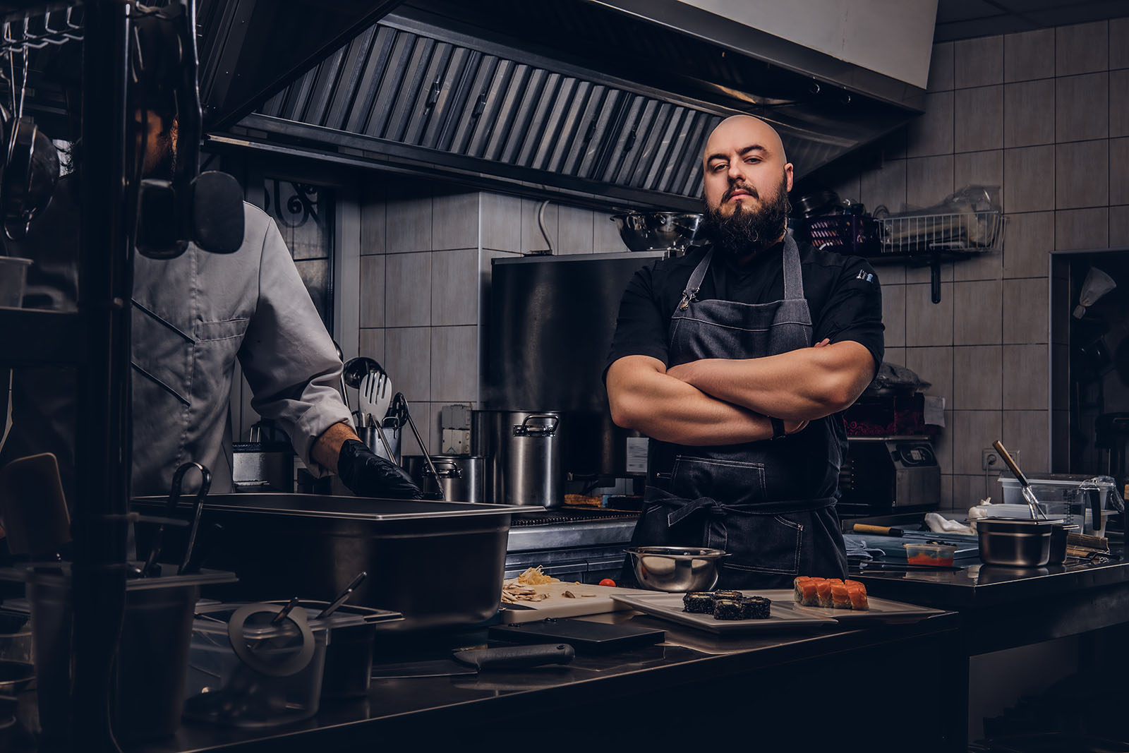 Two bearded cooks dressed in uniforms preparing sushi in a kitchen.